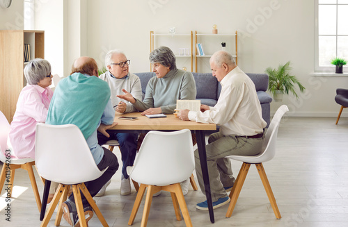 Group of senior people men and women talking with each other, reading a book in nursing home sitting at the table. Pensioners spending leisure time together. Leisure in retirement home concept.