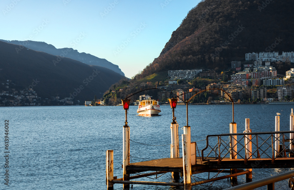 Lake Lugano: at dawn on a clear day, the pier poles on the calm water frame an approaching approaching tourist ferry. in the background rise the mountains