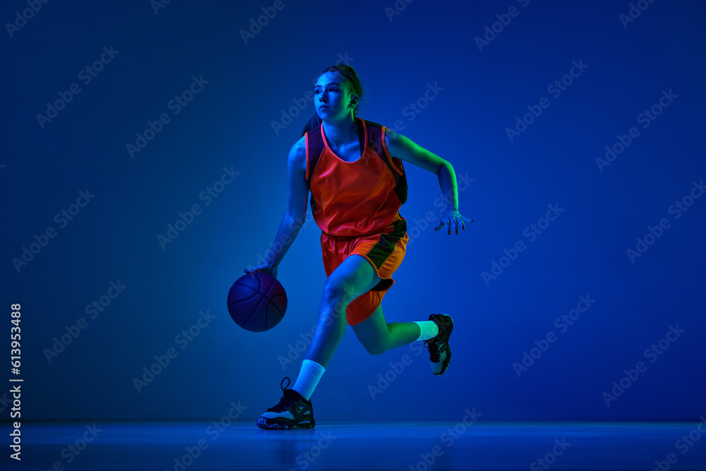Concentrated female basketball athlete training, dribbling ball against blue studio background in neon light. Concept of professional sport, action and motion, game, competition, hobby, ad