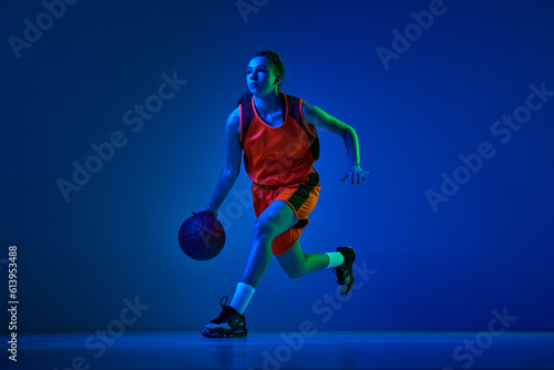 Concentrated female basketball athlete training, dribbling ball against blue studio background in neon light. Concept of professional sport, action and motion, game, competition, hobby, ad © master1305