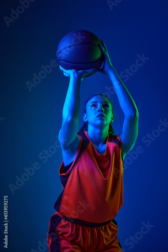 Winning goal. Young girl, concentrated basketball player throwing ball against blue studio background in neon light. Concept of professional sport, action and motion, game, competition, hobby, ad