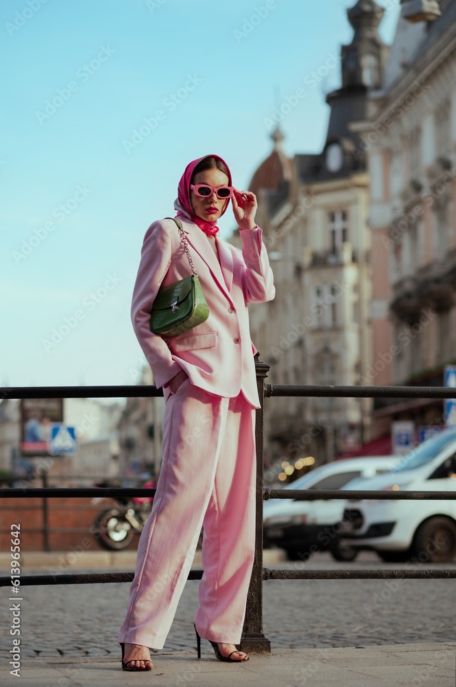 Fashionable elegant confident woman wearing trendy pink sunglasses, silk headscarf, suit blazer, wide leg trousers, with green bag, posing in street of European city. Outdoor full-length portrait