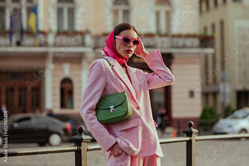 Fashionable elegant confident woman wearing trendy pink sunglasses, silk headscarf, suit blazer, with green bag, posing in street of European city. Outdoor fashion portrait. Copy, empty space for text