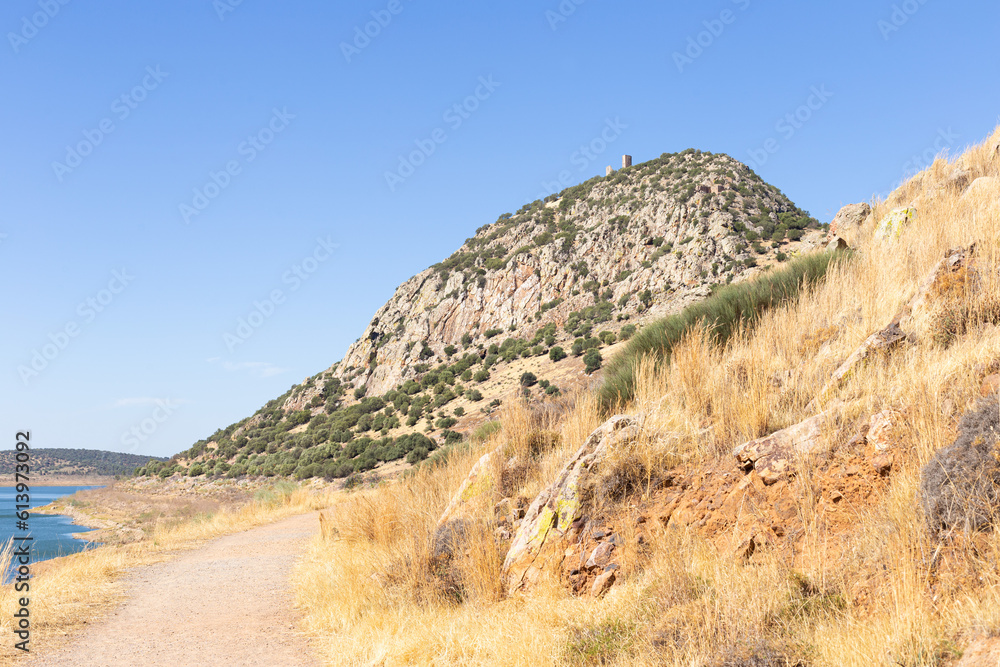 Dirt road that goes to small mountain that has tower above. There is lake on left side and hillside with dry grass on its right. Sky is blue. Alange, Extremadura, Spain.