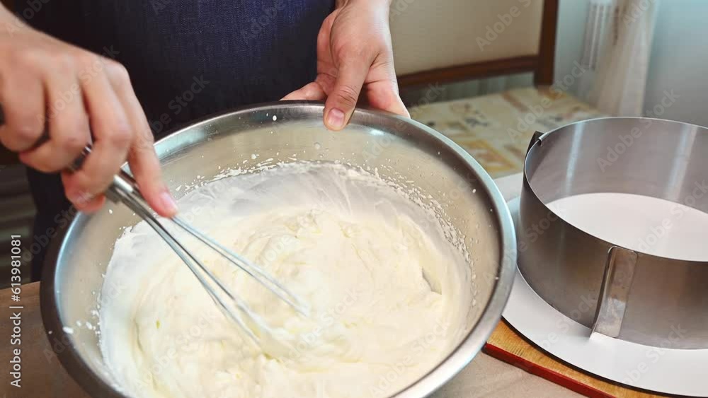 Close-up of a pastry chef using a whisk, whipping fresh cream and mixing with powdered sugar in a metal bowl. preparing a mousse cake in the kitchen. Confectionery. Bakery. Cooking. Food industry Close-up of a pastry chef using a whisk, whipping fresh cream and mixing with powdered sugar in a metal bowl. preparing a mousse cake in the kitchen. Confectionery. Bakery. Cooking. Food industry