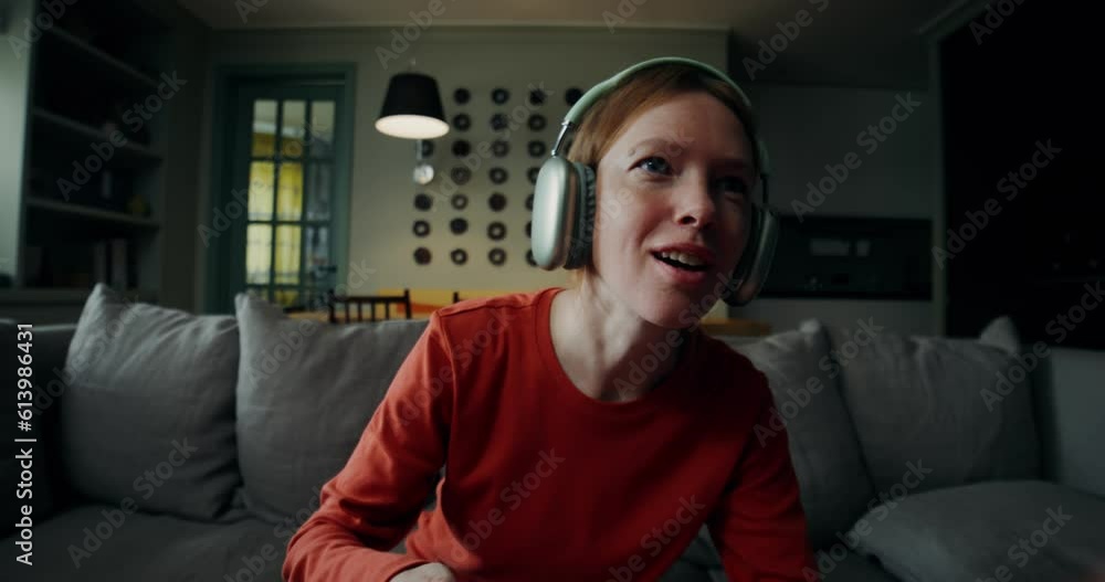 A young woman in casual clothes, wearing headphones, smiling playing video games with a joystick, sitting on the couch at home A young woman in casual clothes, wearing headphones, smiling playing video games with a joystick, sitting on the couch at home