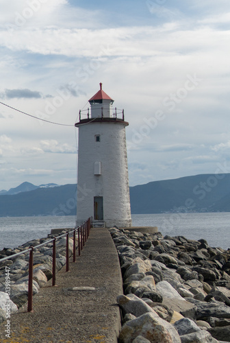 A small lighthouse at the tip of a rocky cape with mountains, fjords in the background and cloudy sky