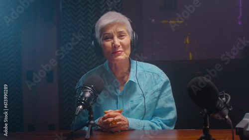 Portrait: Serious old mature blogger woman in wireless headphones recordings podcast in a professional recording studio. An elderly lady on a radio interview. DJ creates live streaming on the Internet photo