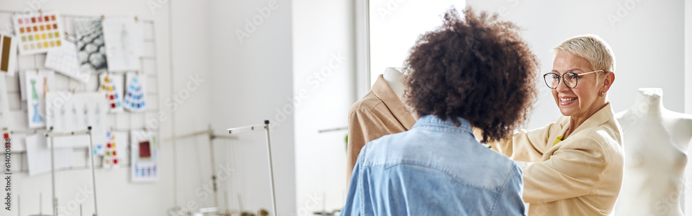 Happy middle aged lady dressmaker teaches young African-American stagist in workshop Happy middle aged lady dressmaker teaches young African-American stagist in workshop
