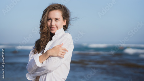 Portrait of a wealthy young woman in a white shirt and blue bikini, standing on the Bali coast of the ocean on a sunny day against the blue sky, smiling and looking. Traveling in the southern Asia.