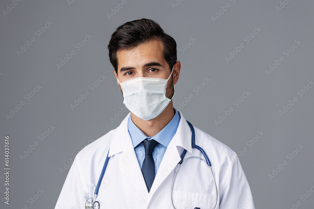 A male doctor in a white coat and medical mask and sterile gloves looks at the camera on a gray isolated background, copy space, space for text