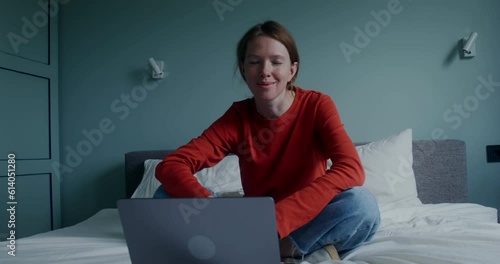 A young red-haired woman smiles looking at the camera while sitting on the bed in front of an open laptop. Stylish home interior photo