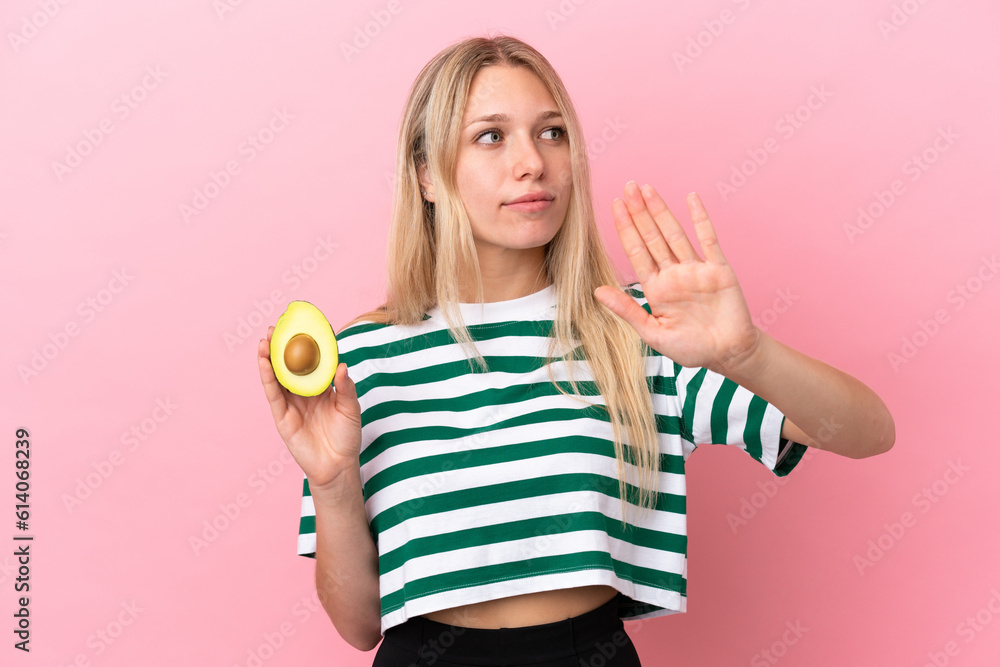 Young caucasian woman holding an avocado isolated on pink background making stop gesture and disappointed Young caucasian woman holding an avocado isolated on pink background making stop gesture and disappointed