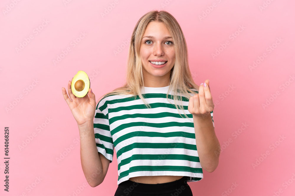 Young caucasian woman holding an avocado isolated on pink background making money gesture Young caucasian woman holding an avocado isolated on pink background making money gesture