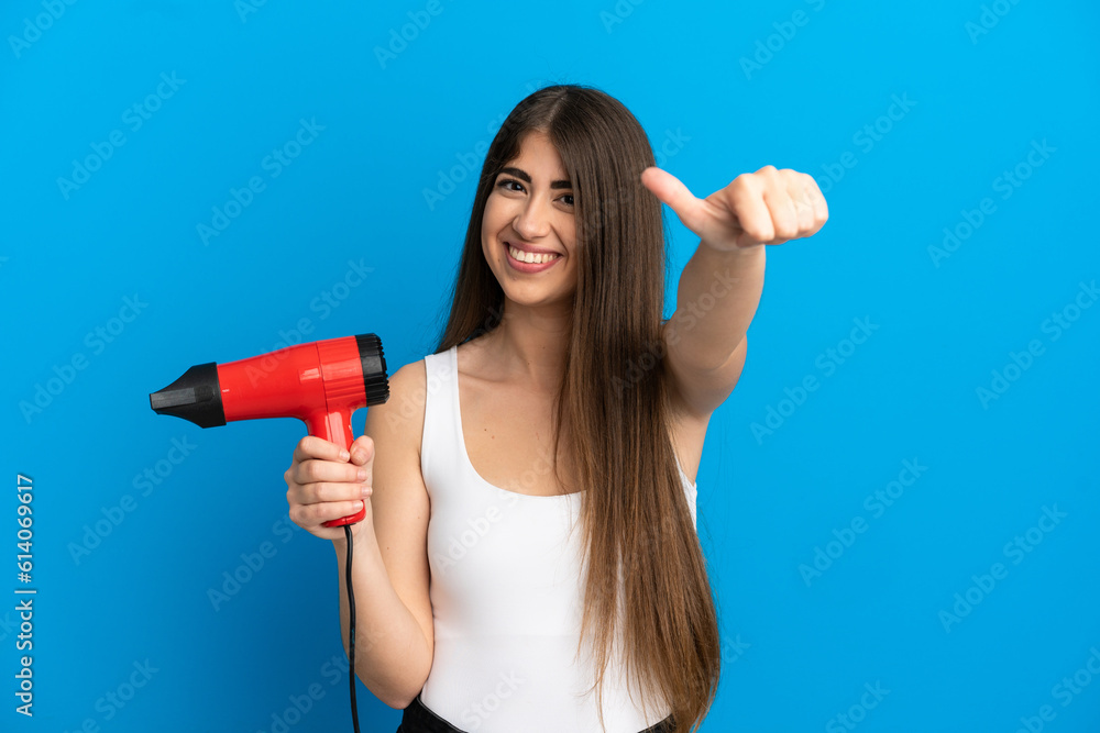 Young caucasian woman holding a hairdryer isolated on blue background giving a thumbs up gesture Young caucasian woman holding a hairdryer isolated on blue background giving a thumbs up gesture