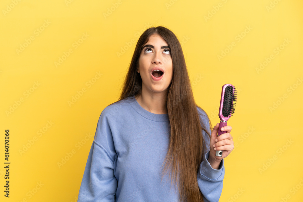 Young caucasian woman holding hairbrush isolated on blue background looking up and with surprised expression Young caucasian woman holding hairbrush isolated on blue background looking up and with surprised expression