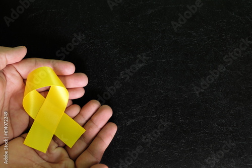 Human hands holding yellow ribbon color on dark black background. Bone cancer awareness and suicide prevention hope concept.