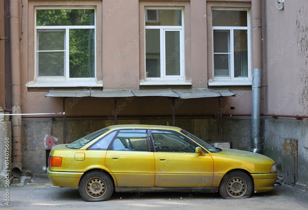 An old yellow car with deflated wheels is parked against the wall of a residential building, Tallinn Street, Saint Petersburg, Russia, June 14, 2023
