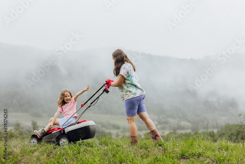A woman in boots with her child in the form of a game mows the grass with a lawnmower in the garden against the background of mountains and fog, garden tools concept