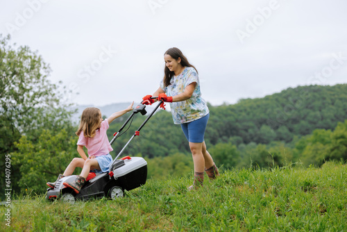 A woman in boots with her child in the form of a game mows the grass with a lawnmower in the garden against the background of mountains and fog, garden tools concept