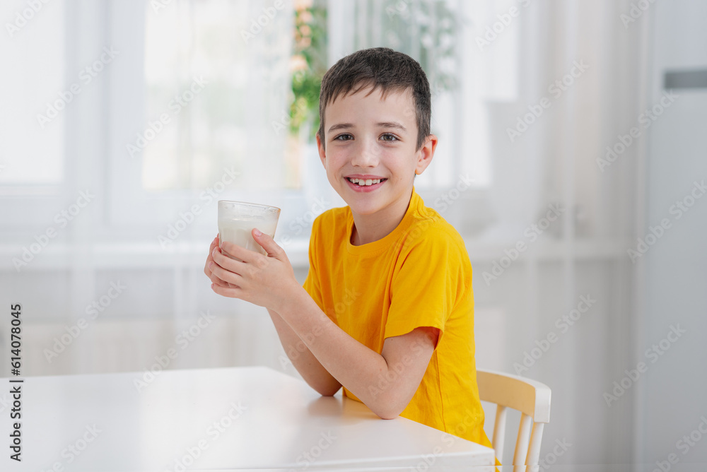 A boy in a bright yellow T-shirt sits at the kitchen table and holds a glass of milk in his hands and smiles broadly at the camera. The benefits of milk for children A boy in a bright yellow T-shirt sits at the kitchen table and holds a glass of milk in his hands and smiles broadly at the camera. The benefits of milk for children