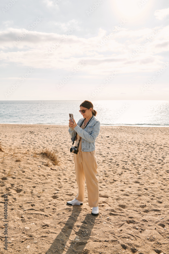 A young, dark, mature woman has an old camera around her neck while taking a picture with her phone