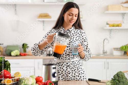Young woman pouring vegetable juice from blender into glass in kitchen