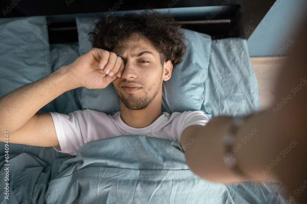 Top view of handsome man 20s with dark hair taking selfie photo while lying in bed under white linen, rubbing his eyes Top view of handsome man 20s with dark hair taking selfie photo while lying in bed under white linen, rubbing his eyes