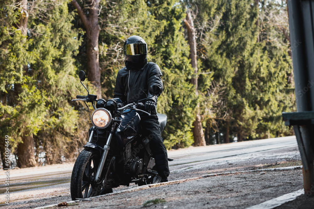 motorcyclist in a leather jacket and helmet on a custom motorcycle cafe racer on a forest road