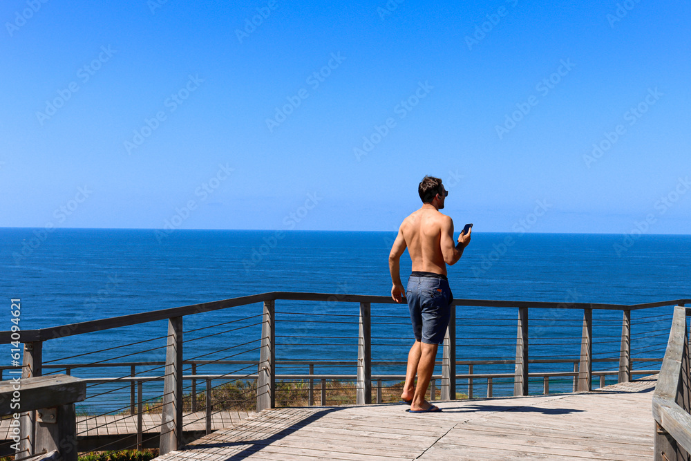 Young man traveler with his phone in his hands speaking in his headphones in a peaceful place in front of the ocean viewed from the back. Portugal Solo traveler.