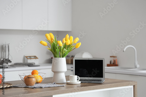 Fototapeta Naklejka Na Ścianę i Meble -  Vase with yellow tulip flowers, modern laptop, fruits and cup of coffee on wooden table in light kitchen