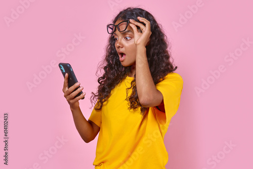Young surprised Indian woman with phone raises glasses from eyes after seeing unexpected message from ex-boyfriend or reading news in yellow press stands on isolated pink background. photo