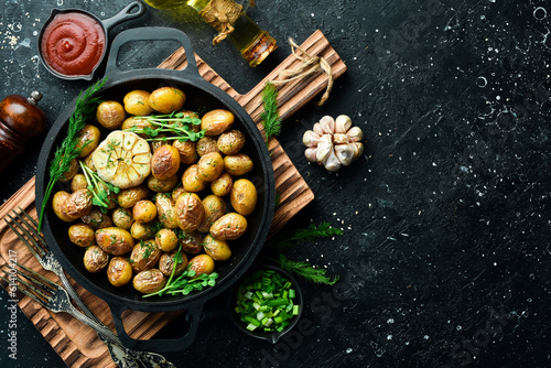 Fototapeta Naklejka Na Ścianę i Meble -  Appetizing baked potatoes whole in a pan. Rustic baked potato with garlic, herbs and spices. On a black stone background.