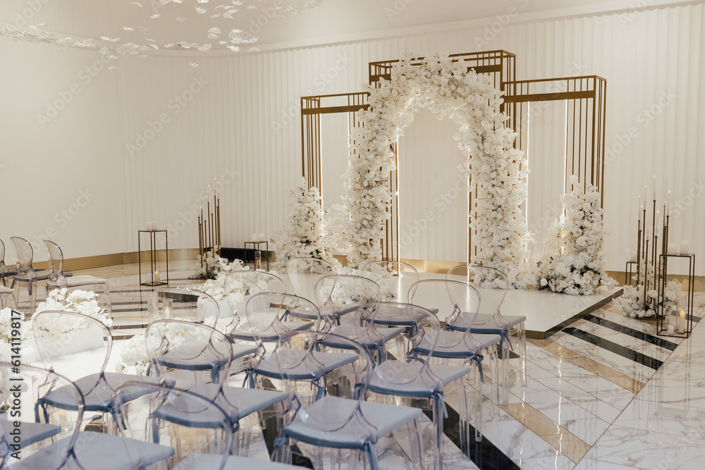 Beautiful modern wedding arch for unique luxury wedding ceremony with white fresh flowers. Transparent chairs near.