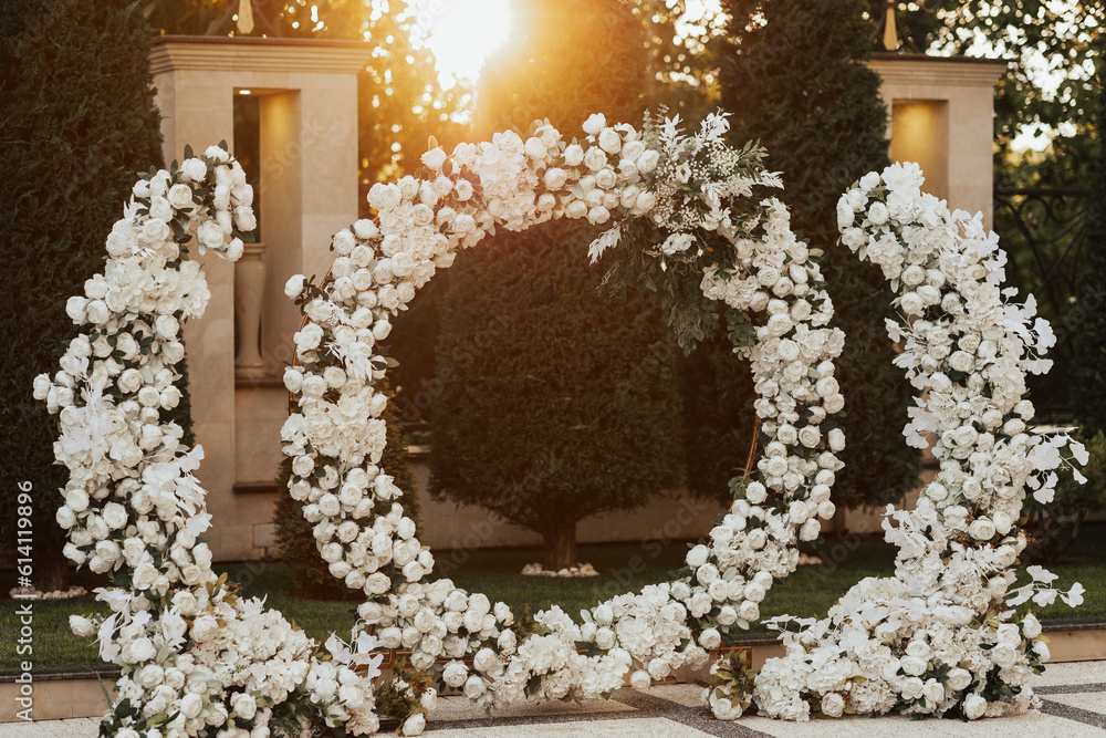 Round arch decorated with white flowers and greenery in the garden at sunset. Area of the wedding ceremony. Beautiful wedding set up.
