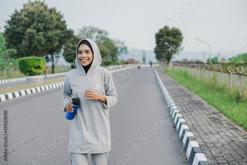 asian muslim woman running and exercising while holding drink bottle outdoors excitedly © rizkiwanggono