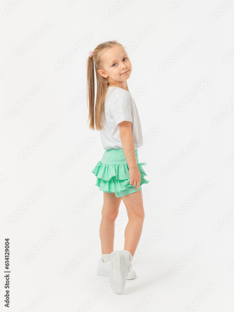 A 5-year-old girl with long hair is standing sideways in a T-shirt and holding on to her skirt, looking at the camera in sneakers on a white background. A 5-year-old girl with long hair is standing sideways in a T-shirt and holding on to her skirt, looking at the camera in sneakers on a white background.
