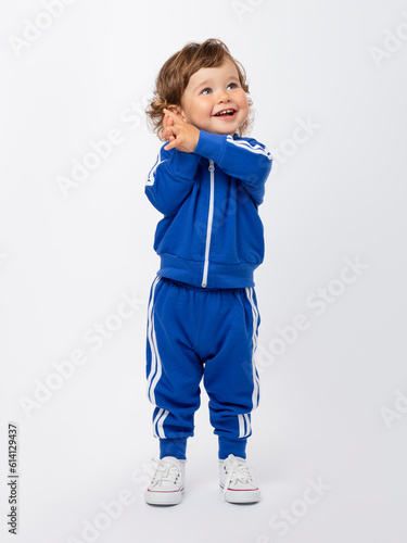A laughing toddler boy of 1-2 years old stands, smiles and dreamily looks up in a blue tracksuit and sneakers on a white background.
