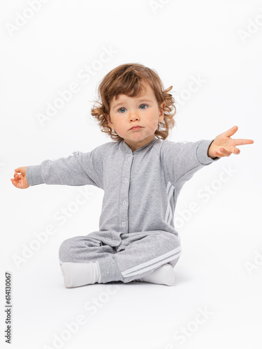 Surprise, question, disappointment. A 2-year-old toddler with curly hair in a gray jumpsuit and socks is sitting on the floor, arms outstretched and legs crossed on a white background.