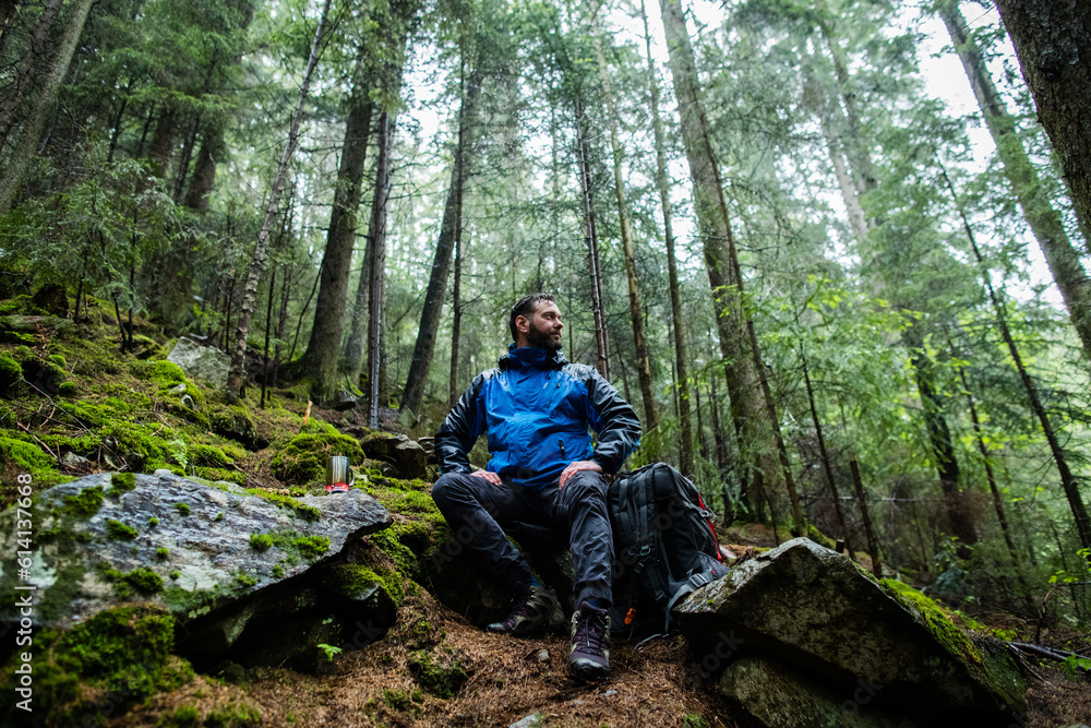 A man-traveler sits on a stone in the forest, a hot drink is being prepared on a gas burner next to him. It is raining in the forest. Solo trips to wild mountain places. A man-traveler sits on a stone in the forest, a hot drink is being prepared on a gas burner next to him. It is raining in the forest. Solo trips to wild mountain places.