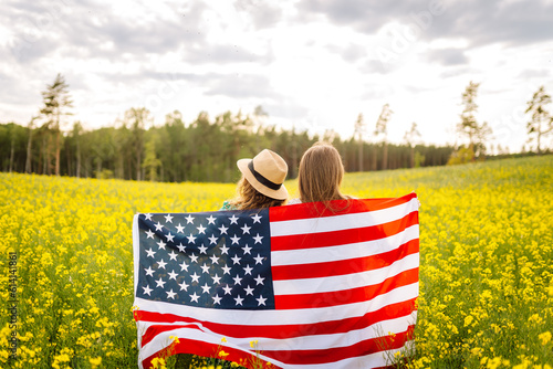 Two Young woman with american flag on blooming meadow. 4th of July. Independence Day. Patriotic holiday. USA flag fluttering in the wind.