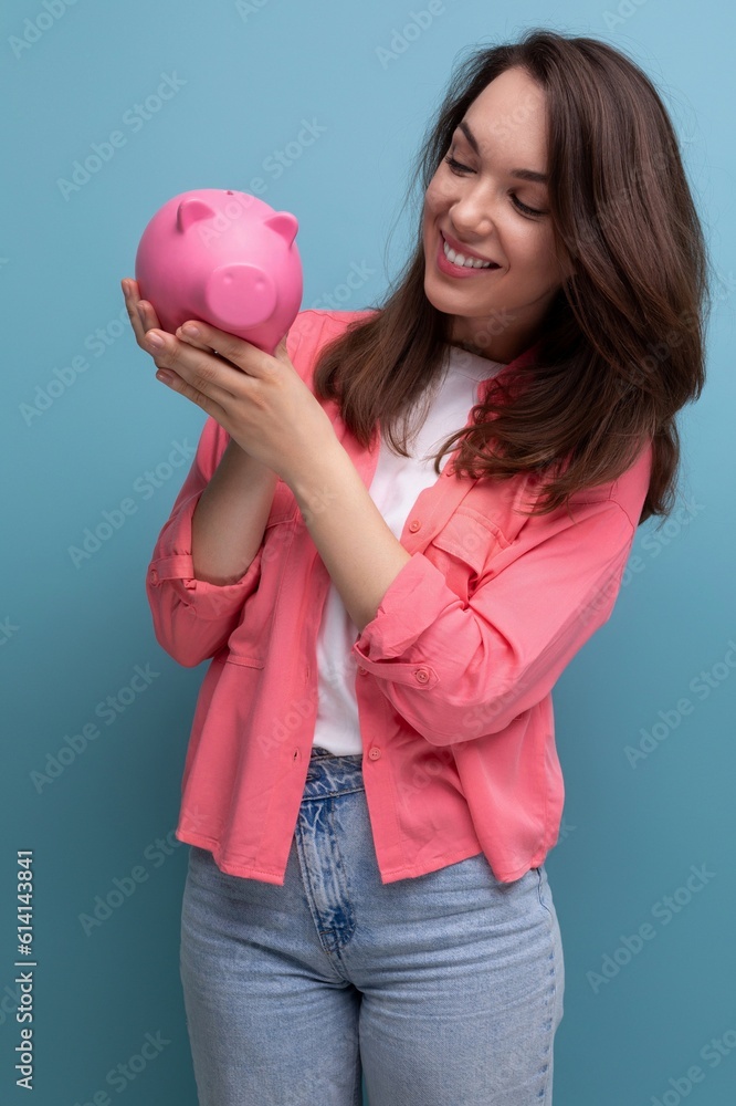 positive brunette young woman in a shirt and jeans puts her budget into a piggy bank positive brunette young woman in a shirt and jeans puts her budget into a piggy bank