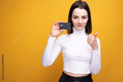 Close up portrait of young smiling business woman holding credit card, isolated on yellow background.
