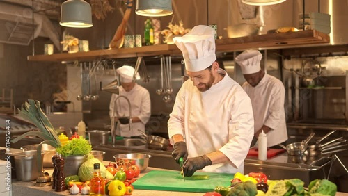 Smiling cook in uniform shredding green fresh cucumber for prepearing salad with sharp kitchen knife. Joyful chef cooking delicious food at professional restaurant kitchen.