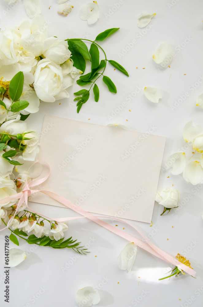 Delicate composition of blank card with white roses and petals with acacia flowers and pink ribbon on white background. Wedding concept. Invitation card template