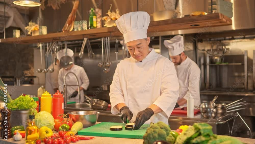 Portrait of asian male chef cutting vegetables in modern kitchen. Professional multicultural cook chopping eggplant on table. Preparation of vegetarian dish with fresh vegetables Portrait of asian male chef cutting vegetables in modern kitchen. Professional multicultural cook chopping eggplant on table. Preparation of vegetarian dish with fresh vegetables