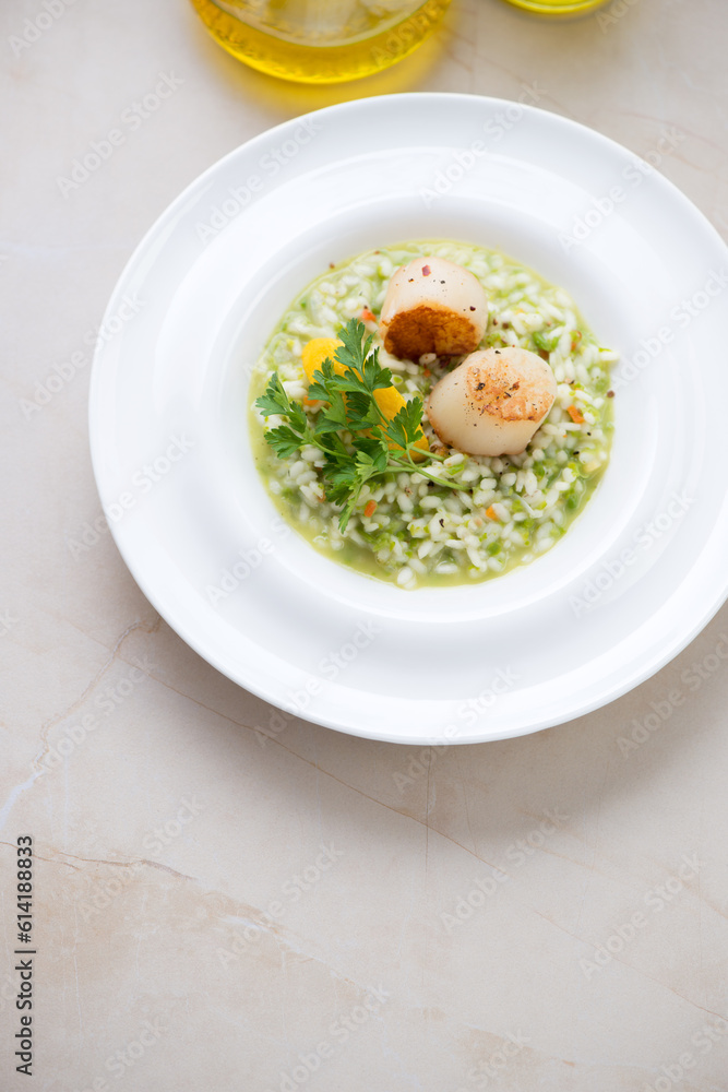 White plate with pea risotto and seared sea scallops, vertical shot on a beige marble background, elevated view White plate with pea risotto and seared sea scallops, vertical shot on a beige marble background, elevated view