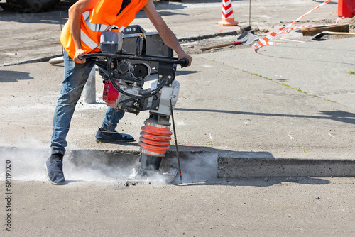 A construction worker is ramming a trench with a vibrating rammer at a construction site and kicking up a cloud of dust around him.