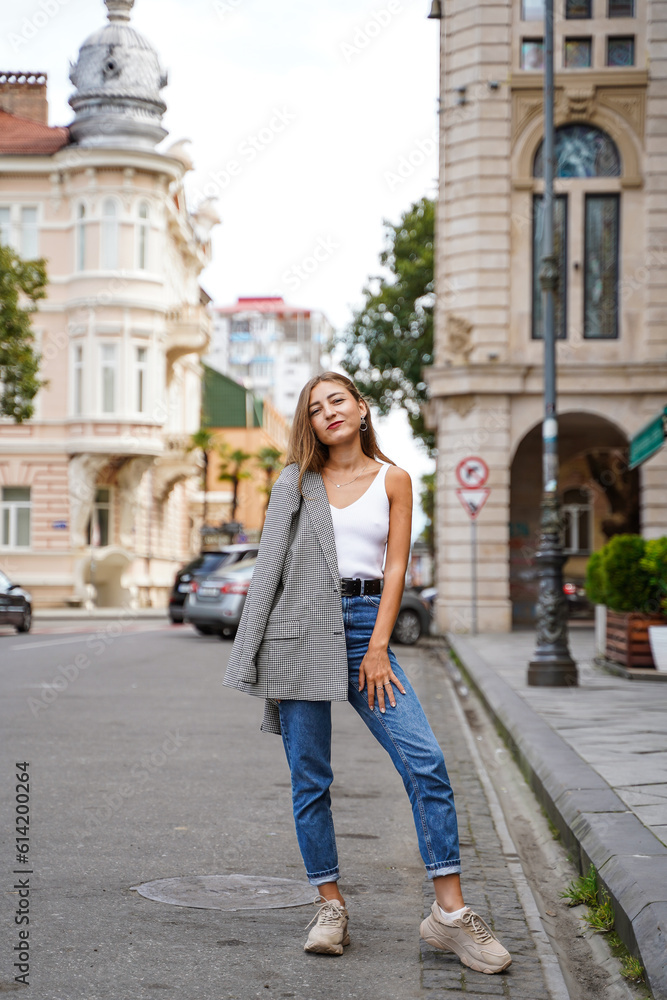 Girl threw jacket on her shoulder. Beautiful young woman wearing casual clothes standing on sidewalk. Historic building facade on background. City recreation. Person resting experiencing calmness