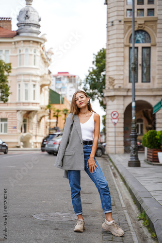 Girl threw jacket on her shoulder. Beautiful young woman wearing casual clothes standing on sidewalk. Historic building facade on background. City recreation. Person resting experiencing calmness